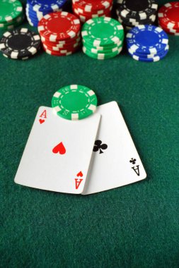 İki kalp ve sinek ası belirgin bir şekilde yeşil bir keçeli masa üzerinde sergilenir, yoğun bir kart oyunu olduğunu gösteren renkli bir poker fişi düzeniyle çevrelenir..