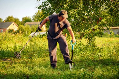Bir adam uzun çimenlerde diz çökerken metal dedektörü kullanıyor. Gün batımında bir tarlada hazine arıyor. Arka planda bir çiftlik evi görülebiliyor ve kırsal atmosfere ekleniyor.