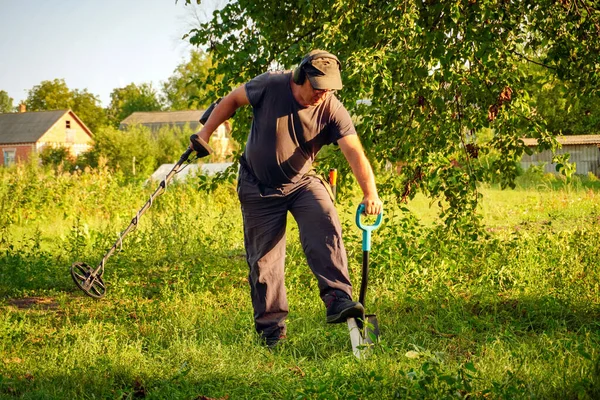 Bir adam uzun çimenlerde diz çökerken metal dedektörü kullanıyor. Gün batımında bir tarlada hazine arıyor. Arka planda bir çiftlik evi görülebiliyor ve kırsal atmosfere ekleniyor.