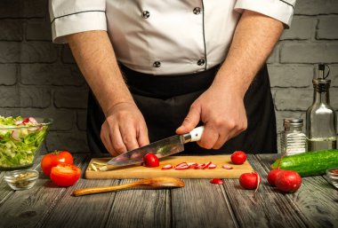 Bir şef ahşap bir kesme tahtasına turp ve domates dilimler. Bölgeyi çevreleyen taze sebzeler, iyi aydınlatılmış bir mutfakta canlı bir aşçılık süreci sergiliyor..