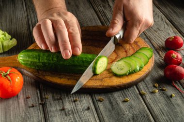 Eller, domates, turp ve yapraklı yeşillikler gibi taze sebzelerle çevrili ahşap bir tahtanın üzerinde ustalıkla salatalık keserek yemek hazırlama becerilerini sergiliyor..