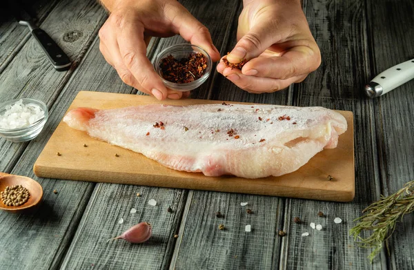 Eller, ahşap kesme tahtasının üzerinde duran çiğ balık filetosunun üzerine baharat serpiyor. Yakınlarda, iyi aydınlatılmış bir mutfak ortamındaki hazırlıklara bir kase tuz ve bütün baharatlar eşlik ediyor..