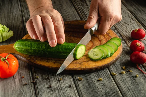 Eller, domates, turp ve yapraklı yeşillikler gibi taze sebzelerle çevrili ahşap bir tahtanın üzerinde ustalıkla salatalık keserek yemek hazırlama becerilerini sergiliyor..