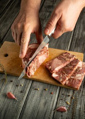 Kırsal bir mutfak ortamında, bir insan çiğ eti tahta bir tahtada dilimlere ayırır. Taze sarımsaklı karanfiller yakınlarda oturur, hazırlık atmosferini arttırır..