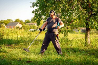 Metal dedektörü kullanan bir adam gizli hazineler için çimenli bölgeyi dikkatlice tarıyor. Parlak güneş ışığı manzarayı aydınlatıyor, ağaçların ve evlerin huzurlu bir kırsal zemin ortaya çıkarıyor..