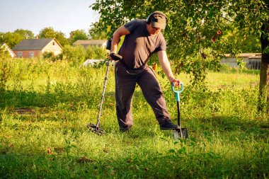 Bir adam öğleden sonra çimenli bir arazide saklı hazineleri aramak için metal dedektör ve kürek kullanıyor. Parlak güneş ışığı çevreyi aydınlatır, canlı yeşili güçlendirir..