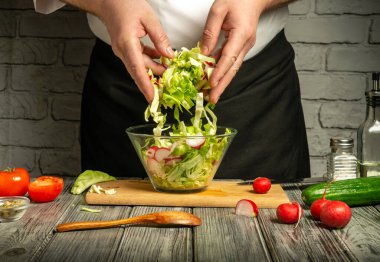 Bir şef ustalıkla taze marul ve turpları temiz bir kasede karıştırır. Onları çevreleyen domatesler ve salatalıklar sıcak bir mutfaktaki tahta kesme tahtasının üzerinde..