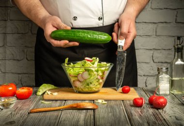 Bir şef, sıcak bir mutfakta taze salata hazırlarken, salatalık da dahil olmak üzere sebzeleri ustalıkla doğrar. Parlak malzemeler masanın üzerinde sergileniyor, tazelik sergileniyor..