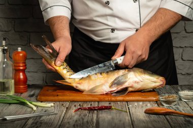 Bir aşçı ustaca fileto yapar ahşap bir tahtanın üzerinde taze bir balık, kırsal bir mutfakta, yemek malzemeleri ve aletlerle çevrili. Hava sıcak ve davetkar..