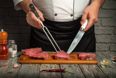 Bir şef sıcak bir mutfakta çatal ve bıçakla ahşap bir tahtada lezzetli biftekler kesiyor. Aromatik baharatlar ve bitkiler etrafa saçılmış, mutfak atmosferini güçlendiriyor..