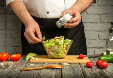 Bir şef, içinde salata, turp, domates ve salatalık bulunan taze hazırlanmış salataya tuz ekler. Mutfağın ahşap yüzeyleri ve tuğla duvarlarıyla kırsal bir havası var..