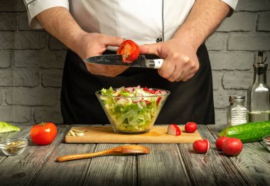 Bir şef marul ve turp kasesinin yanında dururken ustalıkla bir domates keser. Taze salatalıklar ve domatesler yakındaki ahşap bir masanın üzerinde sıcak bir mutfak ortamında..