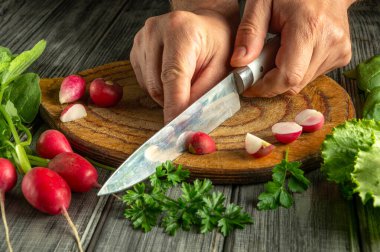 Renkli ürünlerle çevrili ahşap bir kesme tahtasının üzerinde taze turpları ve otları ustaca doğrayan eller. Bu yemek pişirme aktivitesi sağlıklı malzemelerin hazırlanmasını vurguluyor..