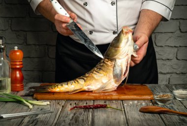 Bir şef usta bir şekilde bütün balığı ahşap bir kesme tahtasının üzerinde doğruyor. Bitki ve baharat gibi taze malzemeler yakınlarda, canlı bir mutfak ortamı sergiliyorlar..