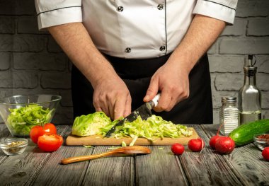 Bir şef lahanayı dilimler ve taze sebzeleri mutfaktaki tahta kesme tahtasında hazırlar. Çeşitli malzemeler arasında domates, salatalık ve salata sosu vardır..