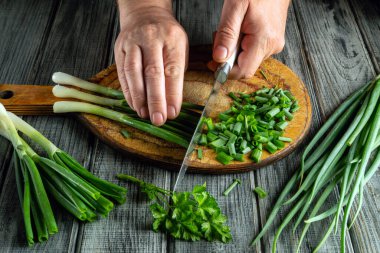 Eller, ahşap bir tahtanın üzerinde yeşil soğan ve taze otları ustalıkla kesiyor. Sahne, doğal ışıklandırmayla sıcak bir mutfakta yemek pişirmenin özünü yakalıyor..