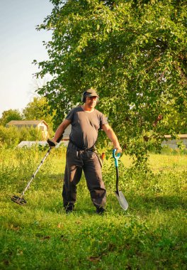 Bir elinde metal detektör, diğer elinde kürek tutarak çimen tarlasında duran şapkalı bir adam. O, bir ağacın yakınında saklı hazineleri ararken güneş sıcacık bir parıltı saçar..