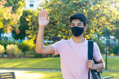 Tıbbi maskeli ve sırt çantalı genç bir adam parkta birine el sallıyor. Coronavirus salgını öğrencisi..