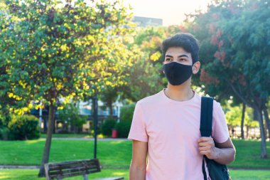 Parkta çantasını tutan tıbbi maske takan genç bir adam. Coronavirus salgını öğrencisi..