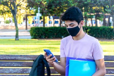 Parktaki bankta oturan tıbbi maskeli genç bir adam telefonunu kullanıyor. Koronavirüs salgını öğrencisi..