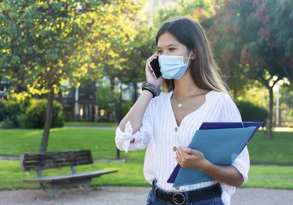 Tıbbi maske takan genç bir kadın telefonla arıyor ve parkta klasörleri tutuyor..