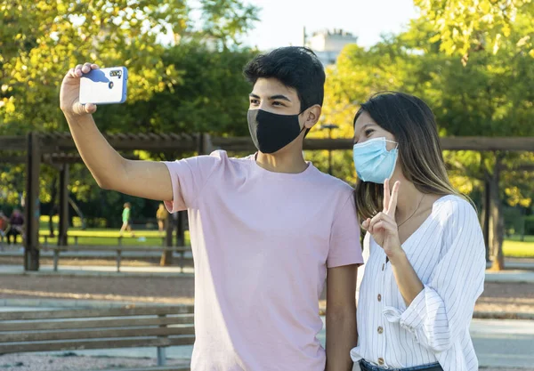 Yüz maskeli kız ve oğlan parkta akıllı telefonuyla selfie çekiyorlar. Yeni normalde arkadaşlık.