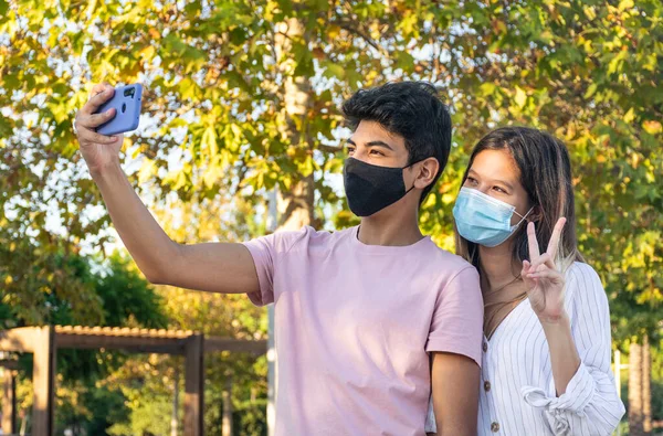 Yüz maskeli kız ve oğlan parkta akıllı telefonuyla selfie çekiyorlar. Yeni normalde arkadaşlık.