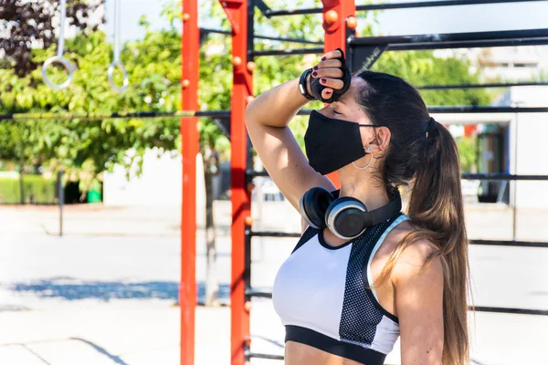 Jimnastik parkında egzersiz ve ısıdan yorgun düşmüş maskeli ve kulaklıklı bir fitness kızı.