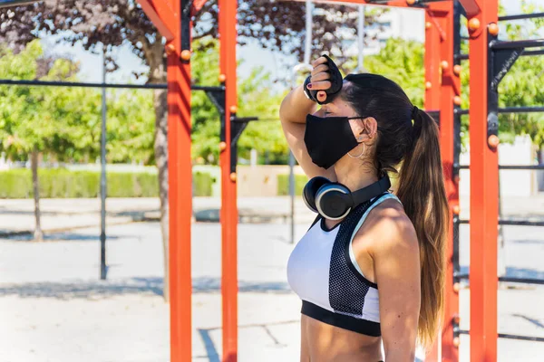 Jimnastik parkında egzersiz ve ısıdan yorgun düşmüş maskeli ve kulaklıklı bir fitness kızı.