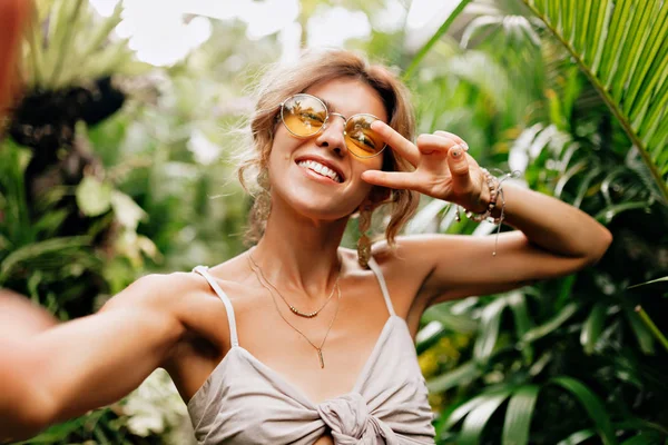 Glückliches Mädchen mit durchtrainierter Haut und weißen Zähnen, das eine gelbe runde Brille trägt, Selfie macht und ein Friedenszeichen in den Tropen zeigt — Stockfoto