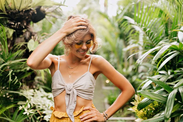 Stilvolle moderne junge Frau mit blonder Lockenfrisur, die Sommerkleidung trägt und an einem sonnigen Sommertag durch die Tropen wandert. — Stockfoto