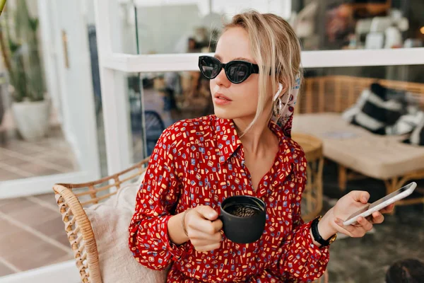 Приваблива стильна біла жінка в чорних сонцезахисних окулярах п'є каву і розмовляє на смартфоні — стокове фото
