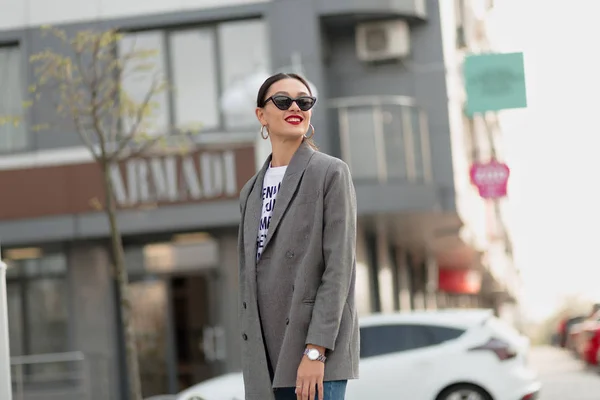 Happy tersenyum wanita berjalan di latar belakang bangunan modern di kota, pusat bisnis. Mengenakan kacamata bergaya, jaket — Stok Foto