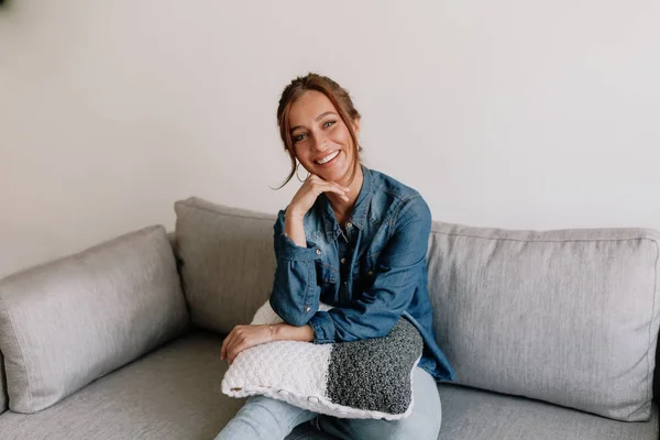 Mujer caucásica sonriente descansando en casa en un acogedor sofá en un apartamento moderno con camisa vaquera y jeans —  Fotos de Stock