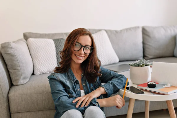 Adorable mujer encantadora con una sonrisa maravillosa usando gafas y camisa de mezclilla trabajando en casa —  Fotos de Stock