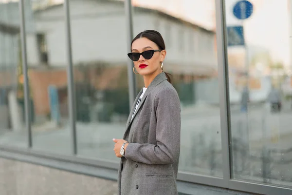 Mujer morena de moda con labios rojos caminando en chaqueta alrededor contra rascacielos. Ella sonriendo a la cámara — Foto de Stock