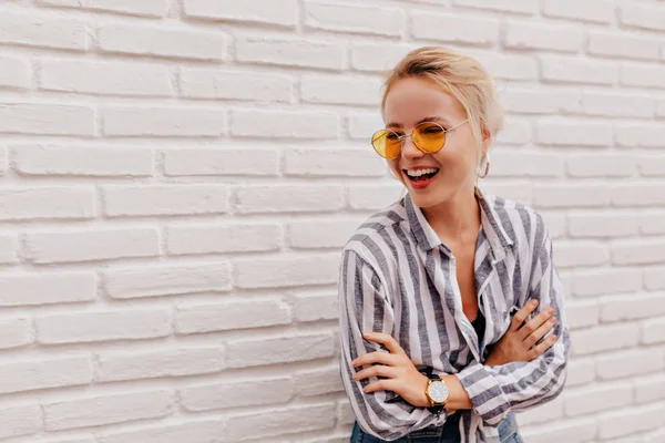 Щаслива чарівна блондинка в стильних помаранчевих окулярах в смугастій сорочці позує з милою посмішкою над білою стіною — стокове фото
