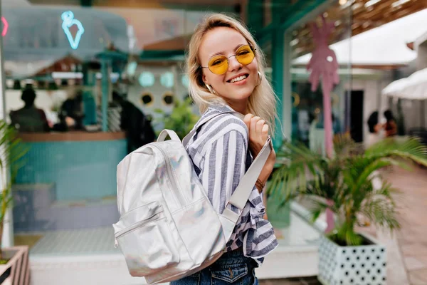 Усміхнена щаслива стильна дівчина в круглих модних спектрах в смугастій сорочці з білим рюкзаком — стокове фото