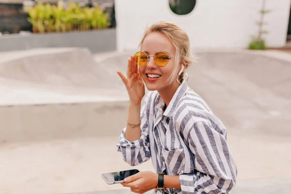 Gadis yang bahagia dengan rambut pirang memakai kacamata oranye mendengarkan musik di headphone dan memegang smartphone — Stok Foto