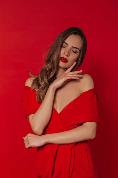 빨간 입술을 입고 빨간 드레스를 입고 눈을 감고 빨간 배경에 포즈를 취하는 매력적인 우아한 여성의 스튜디오 초상화 — 스톡 사진