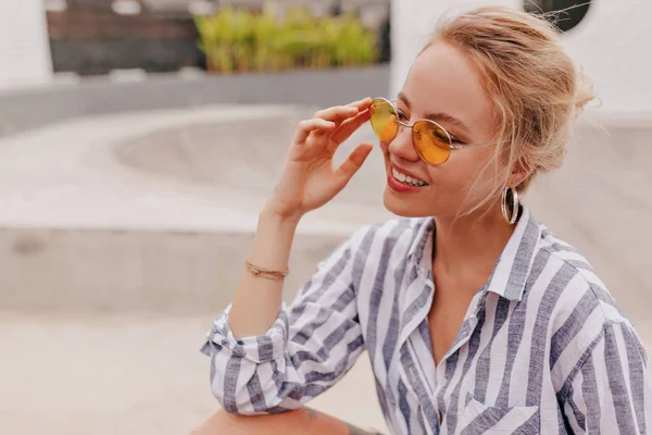 Wanita cantik tersenyum dengan kacamata oranye bundar beristirahat di luar di taman skateboard dan menikmati hari yang panas di musim panas — Stok Foto