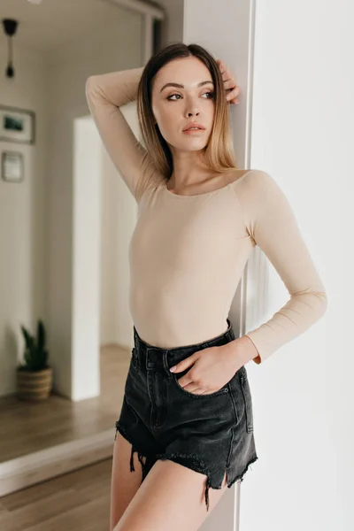 Increíble mujer joven elegante con cuerpo beige y pantalones cortos posando durante la sesión de fotos en el apartamento moderno —  Fotos de Stock