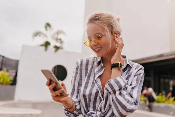 Charming friendly woman in modern round glasses listening music and scrolling smartphone in sunlight