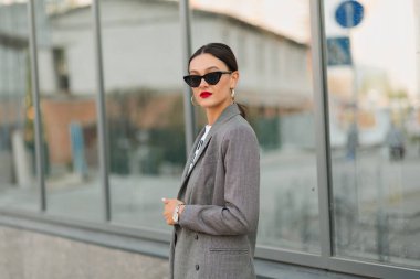 Kırmızı dudakları gökdelen karşı ceket etrafında yürüyüş şık esmer kadın. Kameraya gülümsüyor.