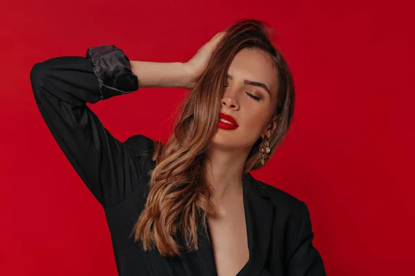 Glamorosa dama con pelo brillante y maquillaje brillante tocando su cabello y ojos cerrados con sonrisa romántica sobre fondo rojo —  Fotos de Stock