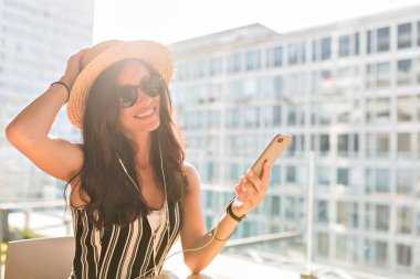 Bulanık kentsel arka planda çatıda akıllı telefon ile oturan hasır şapka koyu saçlı sevimli Avrupalı kadın