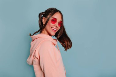 Mavi arka plan üzerinde yuvarlak gözlük takan kameraya gülümseyen koyu saçlı mutlu gülümseyen büyüleyici kız