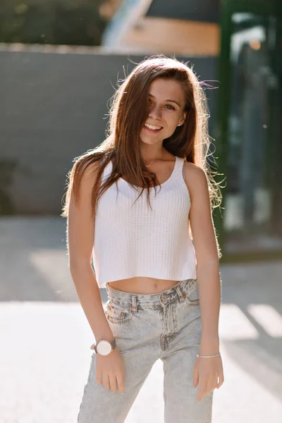 Очаровательная улыбающаяся девушка с длинными волосами в белой футболке и джинсах, гуляющая по городу в солнечный летний день — стоковое фото