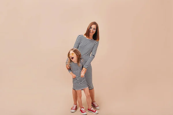 Mãe com 6 anos de idade filha vestindo os mesmos vestidos despojados posando sobre fundo bege com emoções verdadeiras felizes — Fotografia de Stock