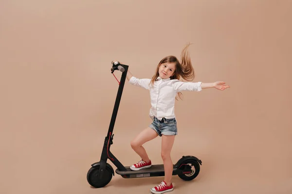 Potret panjang penuh gadis cantik menggemaskan dengan senyum bahagia dan rambut terbang berpose di kamera dengan skuter listrik — Stok Foto